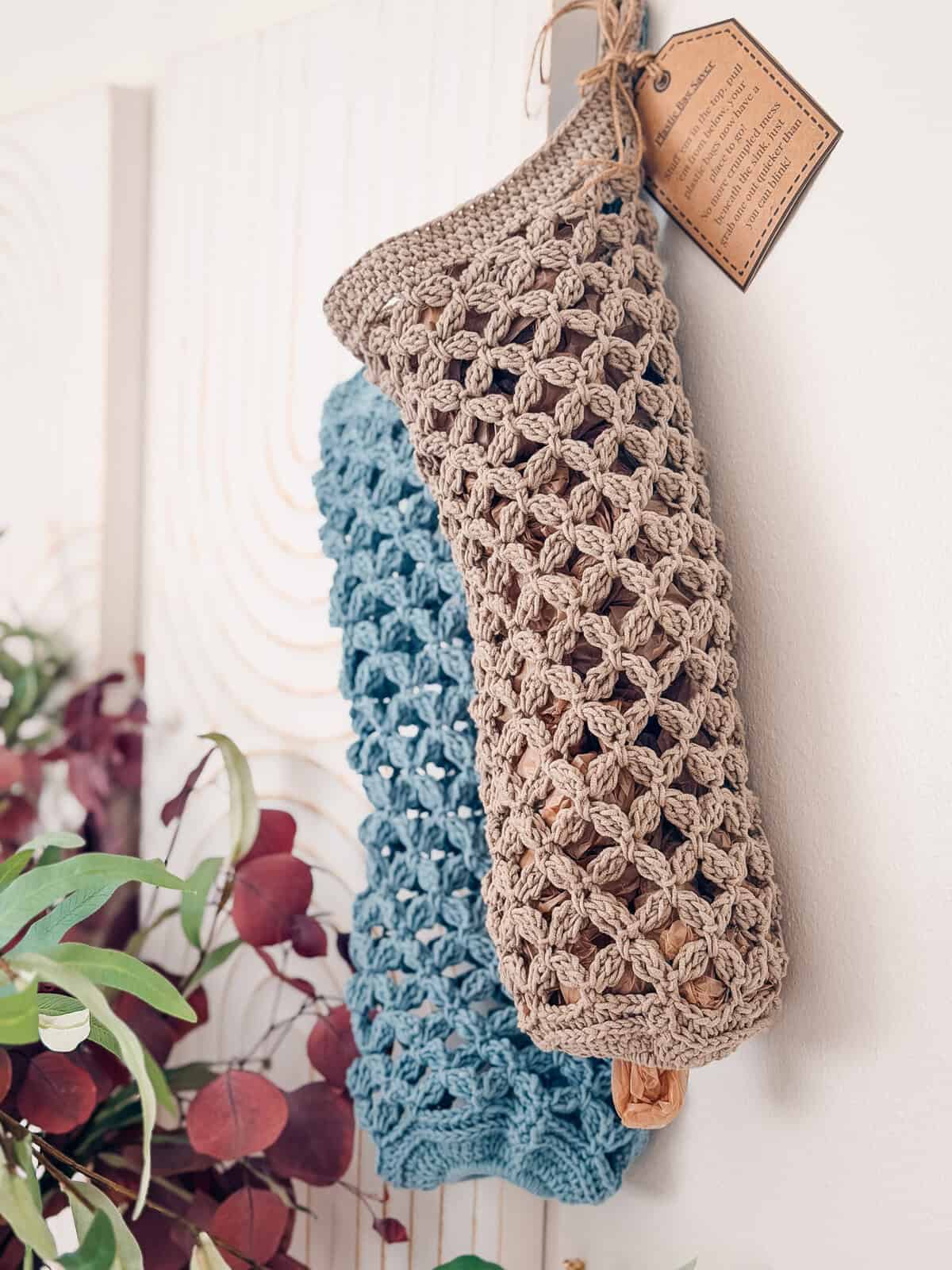 Two crocheted mesh bags, one beige and one blue, hang on a wall. A brown tag is attached to the beige bag. Greenery and purple leaves are visible in the lower left corner.