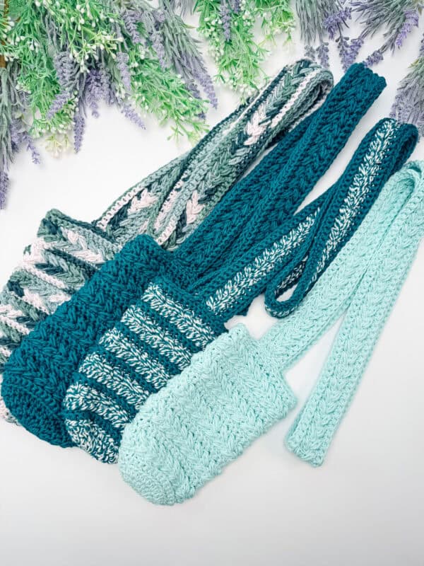 Four crocheted bags in shades of teal and green are arranged side by side on a white surface with greenery and lavender in the background.