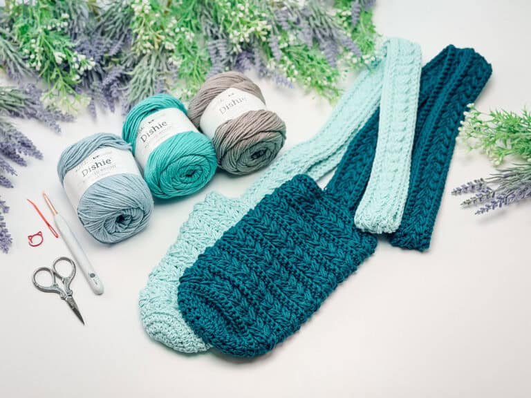 Crocheted teal and mint green bag with matching straps, three balls of yarn, scissors, a hook, and stitch markers on a white surface with greenery in the background.