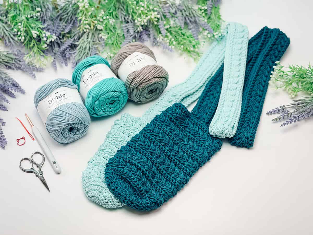 Crocheted teal and mint green bag with matching straps, three balls of yarn, scissors, a hook, and stitch markers on a white surface with greenery in the background.