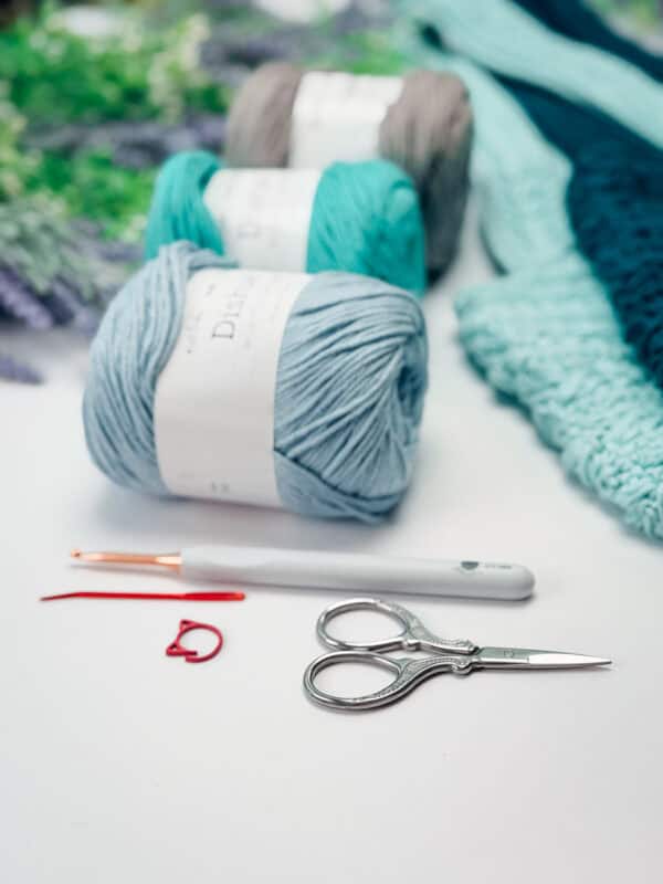 Three skeins of yarn, a crochet hook, a pair of scissors, stitch markers, and a partially completed blue crochet project on a white surface.