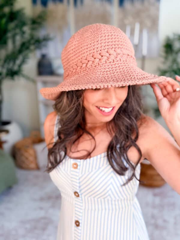 A woman wearing a sleeveless striped dress and a pink woven hat smiles while adjusting the brim, standing indoors with plants and decor in the background.