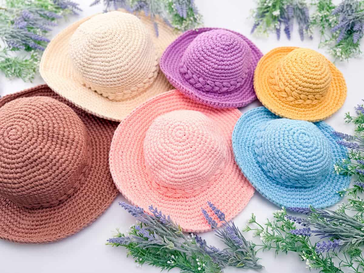 Six crocheted hats in different colors are arranged on a white surface surrounded by sprigs of artificial lavender and greenery.
