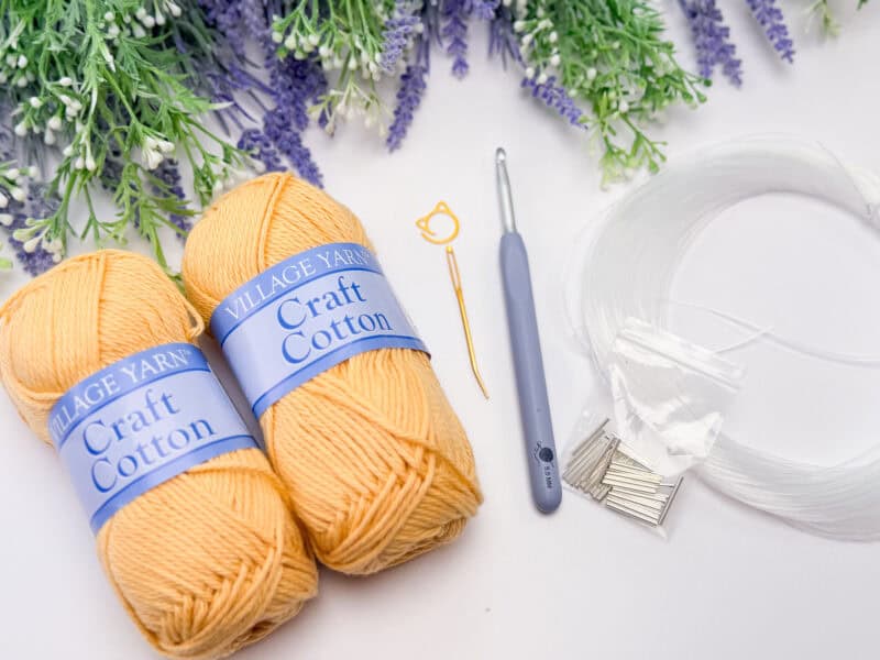Two skeins of yellow craft cotton yarn, a crochet hook, a needle, stitch marker, clear plastic tubing, and metal pins on a white surface with purple and green artificial flowers.
