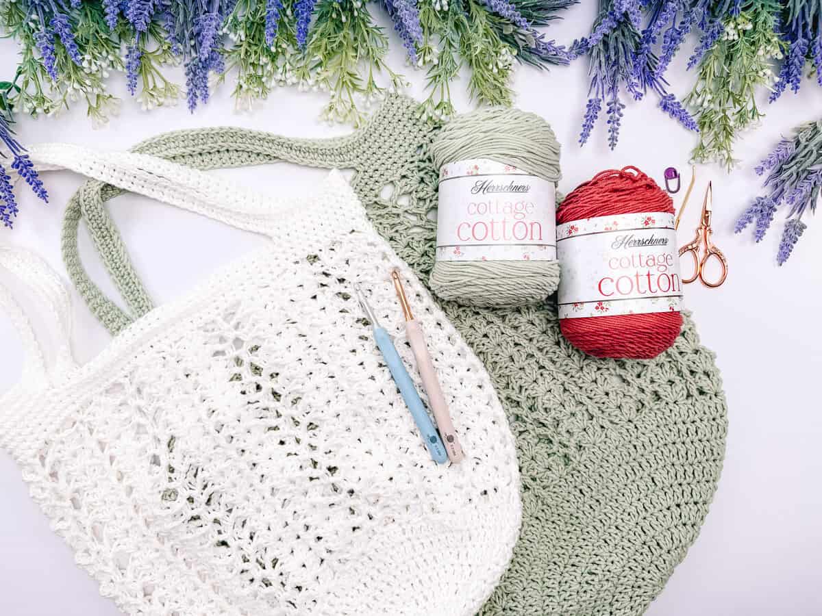 Willow mesh stitch close-up. Crocheted tote bags, two crochet hooks, scissors, and two skeins of cotton yarn are arranged on a white surface with purple flowers around the edges.