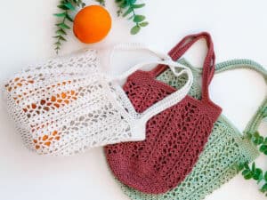 Three crocheted market bags in white, red, and green lie on a white surface with an orange and green leaves nearby.