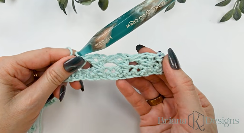 A person holds a partially crocheted light blue swatch with a large crochet hook; the text "Briana K Designs" is visible in the lower right corner.