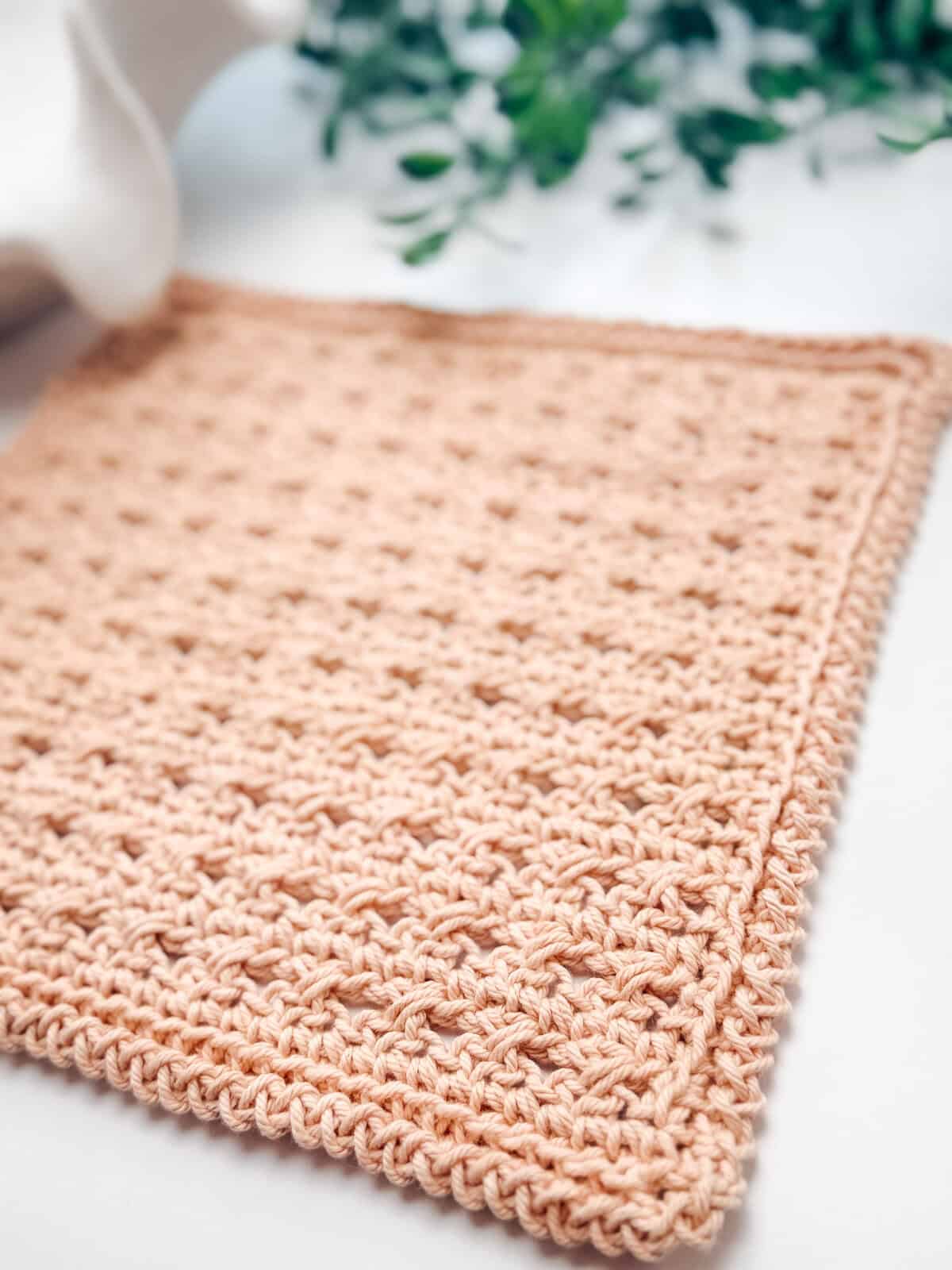 A close-up of a beige crocheted square cloth with a textured pattern, placed on a flat surface with green leaves in the background.