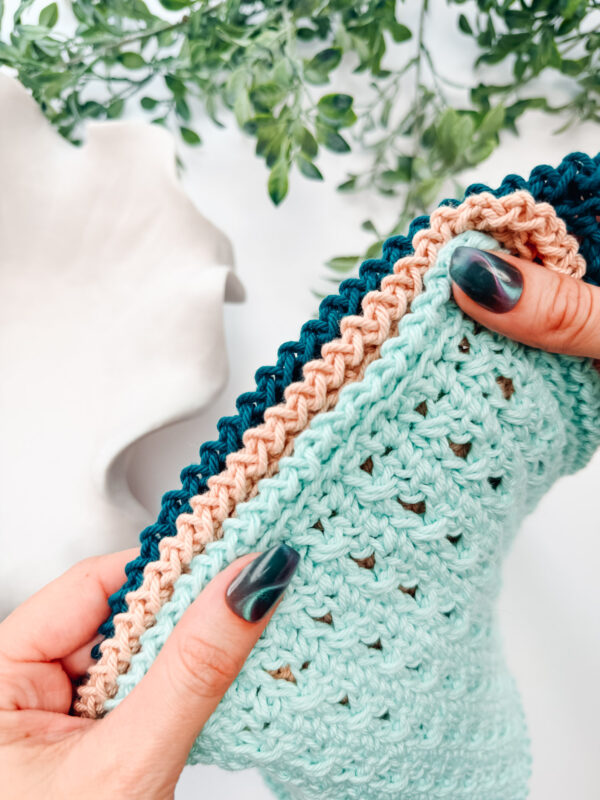 Hands holding a crocheted item with teal, peach, and dark blue edging, against a background of green leaves and a white decorative object.