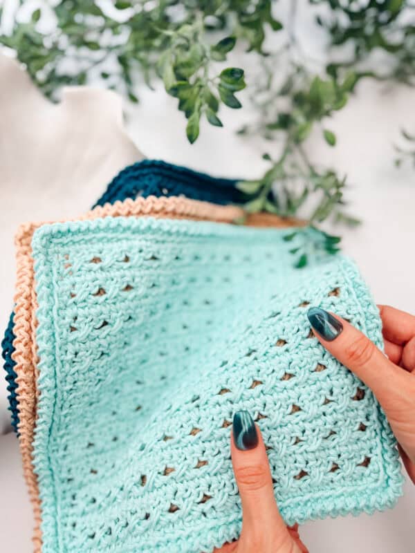 Hands holding a light blue crocheted square with a textured pattern, with other crochet pieces and green foliage in the background.