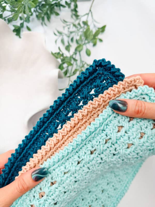 Hands holding neatly folded crocheted cloths in aqua, beige, and dark blue colors, with a white ceramic object and green leafy plant in the background.