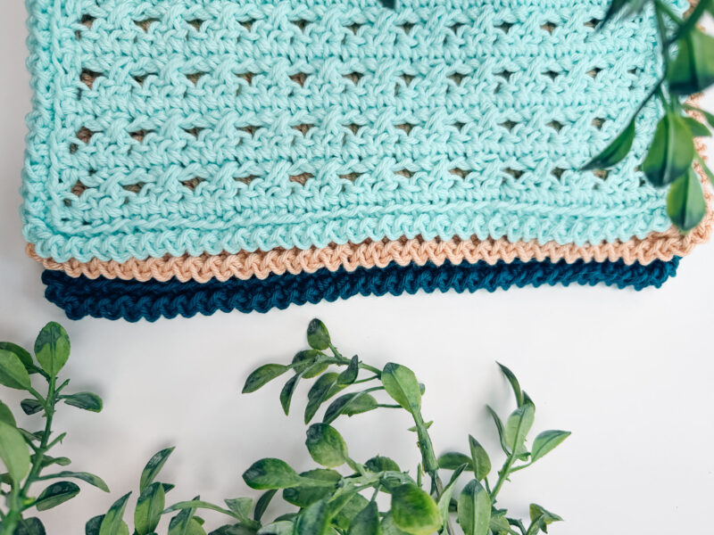 Three folded crochet blankets in mint green, beige, and dark blue are stacked on a white surface with green leafy plants around the edges.