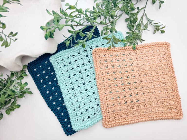 Three crocheted square cloths in navy blue, light blue, and tan are displayed overlapping on a white surface with green leafy branches beside them.