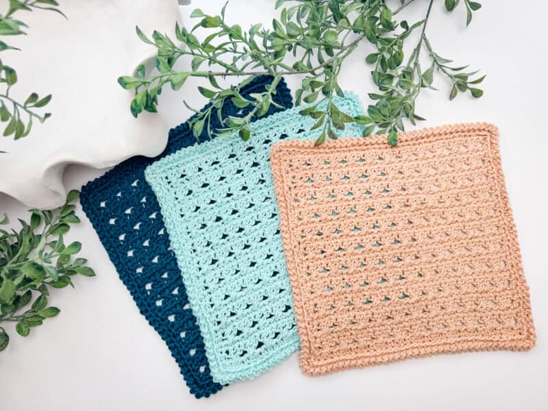 Three crocheted square cloths in navy blue, light blue, and tan are displayed overlapping on a white surface with green leafy branches beside them.
