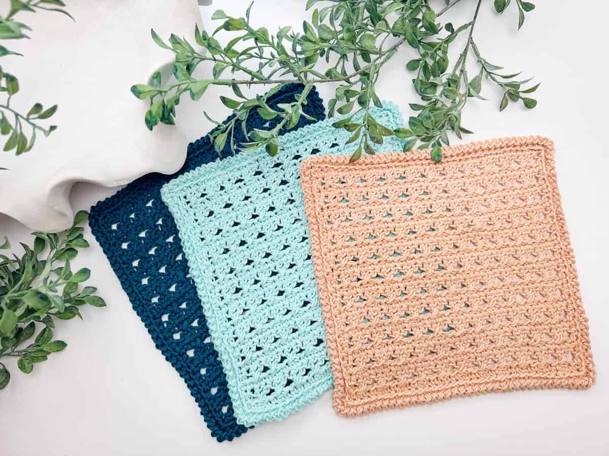Three crocheted square cloths in navy blue, light blue, and tan are displayed overlapping on a white surface with green leafy branches beside them.