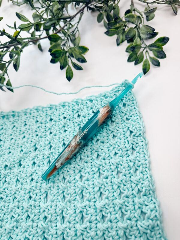 A blue crochet hook rests on a textured aqua crochet fabric, with green leafy branches in the background.
