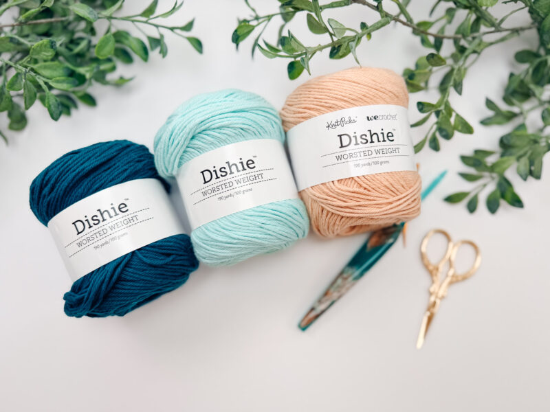 Three balls of Dishie worsted weight yarn in dark blue, light blue, and beige are arranged on a white surface with green leaves, scissors, and a crochet hook.