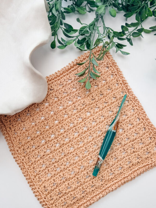A crocheted tan square with a green crochet hook on top, surrounded by green leaves and a white ceramic object on a white surface.