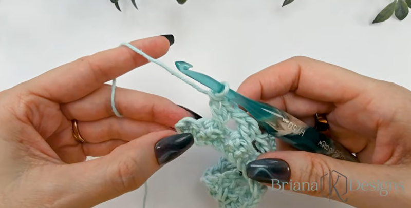 Close-up of hands crocheting with light blue yarn and a teal crochet hook, working on a textured stitch pattern. The watermark reads "Briana K Designs" in the lower right corner.