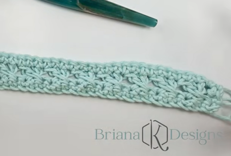 A crocheted light blue swatch with a lacy pattern lies next to a crochet hook. The Briana K Designs logo is visible in the lower right corner.