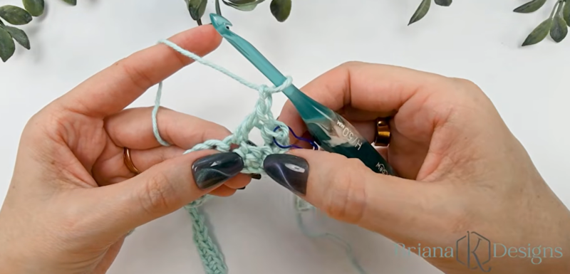 Close-up of hands crocheting with light blue yarn and a teal crochet hook, in the process of making stitches; greenery is visible in the background.