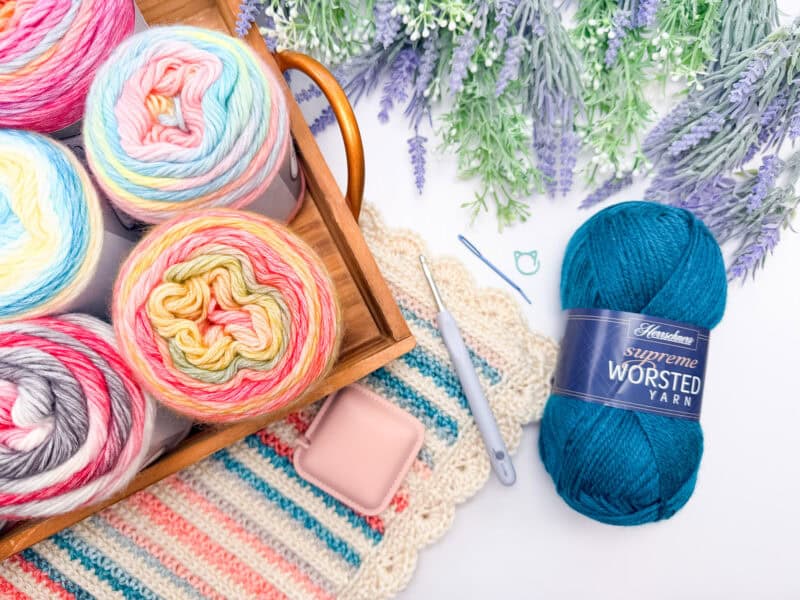 A wooden tray with colorful yarn skeins, a blue ball of worsted yarn, crochet tools, and lavender flowers on a crocheted mat.
