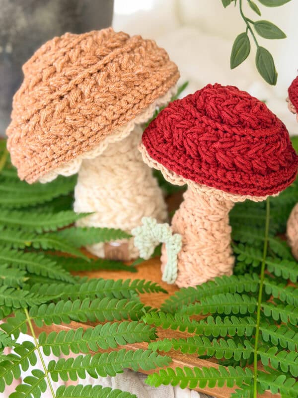 Two crocheted mushrooms, one with a brown cap and one with a red cap, are displayed upright on a wooden surface surrounded by green fern leaves.