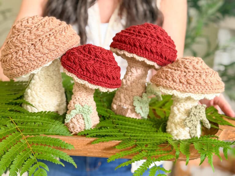 A person holds a wooden tray with crocheted mushrooms in beige and red tones, surrounded by green fern leaves.