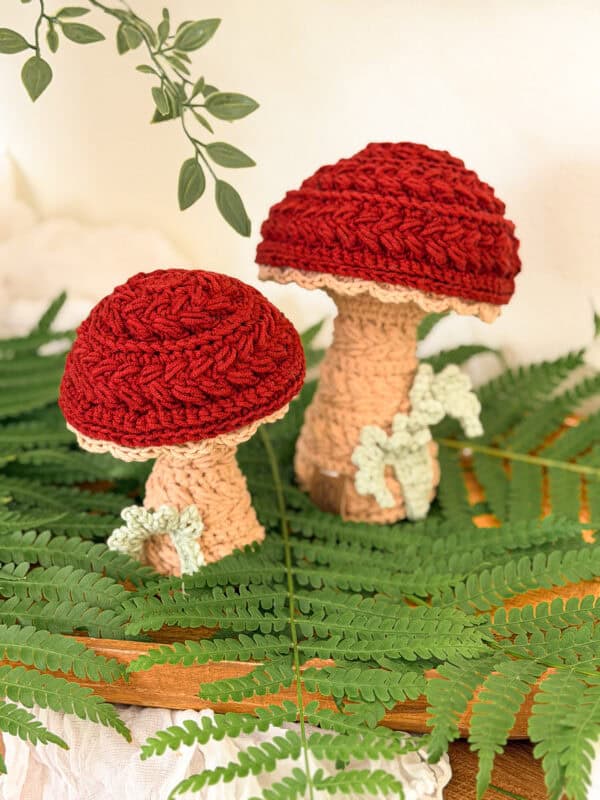 Two crocheted mushrooms with red caps and beige stems are displayed among green ferns and leaves on a wooden surface.