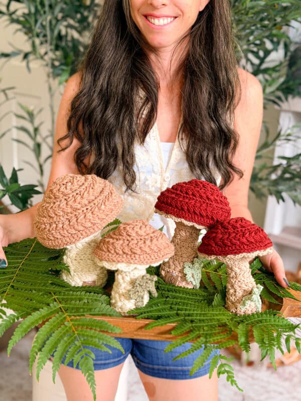 A woman holds a tray with several crocheted mushrooms in different sizes and colors, displayed on green fern leaves.