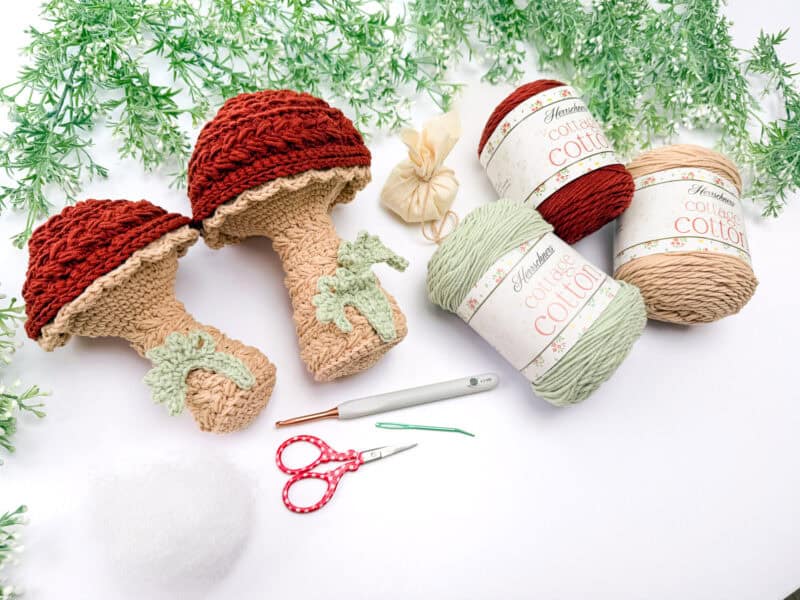 Two crocheted mushroom toys are displayed with yarn balls, stuffing, a crochet hook, scissors, and needles on a white surface, surrounded by artificial greenery.