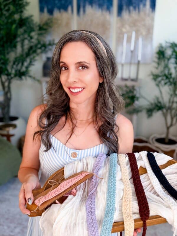 A woman with long brown hair wearing a striped dress holds a wooden rack displaying several colorful braided headbands.