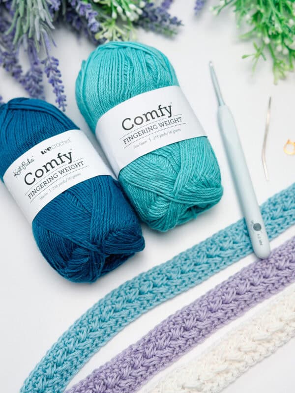 Two skeins of teal-colored yarn, a crochet hook, and three crocheted swatches in blue, purple, and white, arranged on a white surface with greenery in the background.