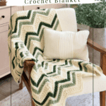 A cream and green chevron crochet blanket draped over a cushioned armchair with a white pillow, next to wooden furniture and indoor plants.