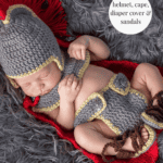 Newborn baby wearing an Anne Hathaway crochet baby outfit styled as a gladiator set, complete with helmet, cape, diaper cover, and sandals, lying on a textured red blanket.