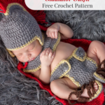 Newborn baby wearing an Anne Hathaway crochet baby outfit styled as a gladiator costume, including helmet, armor, belt, wristbands, and sandals, lying on a red blanket on a gray furry surface.