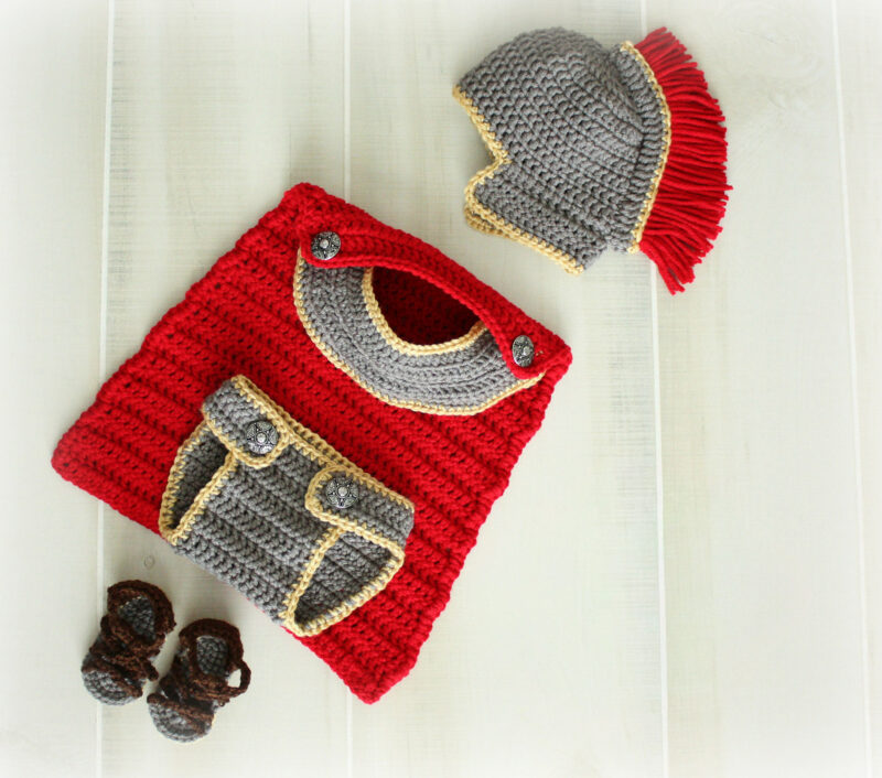 Crocheted baby costume set, inspired by the classic anne hathaway crochet baby outfit style, including a red and gray tunic, gray Roman-style helmet with red crest, and brown sandals, all arranged on a white surface.