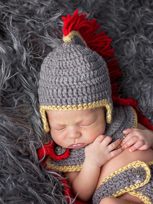 Newborn baby sleeping on faux fur, dressed in an anne hathaway crochet baby outfit styled as a Roman soldier helmet and armor with gold and red accents.