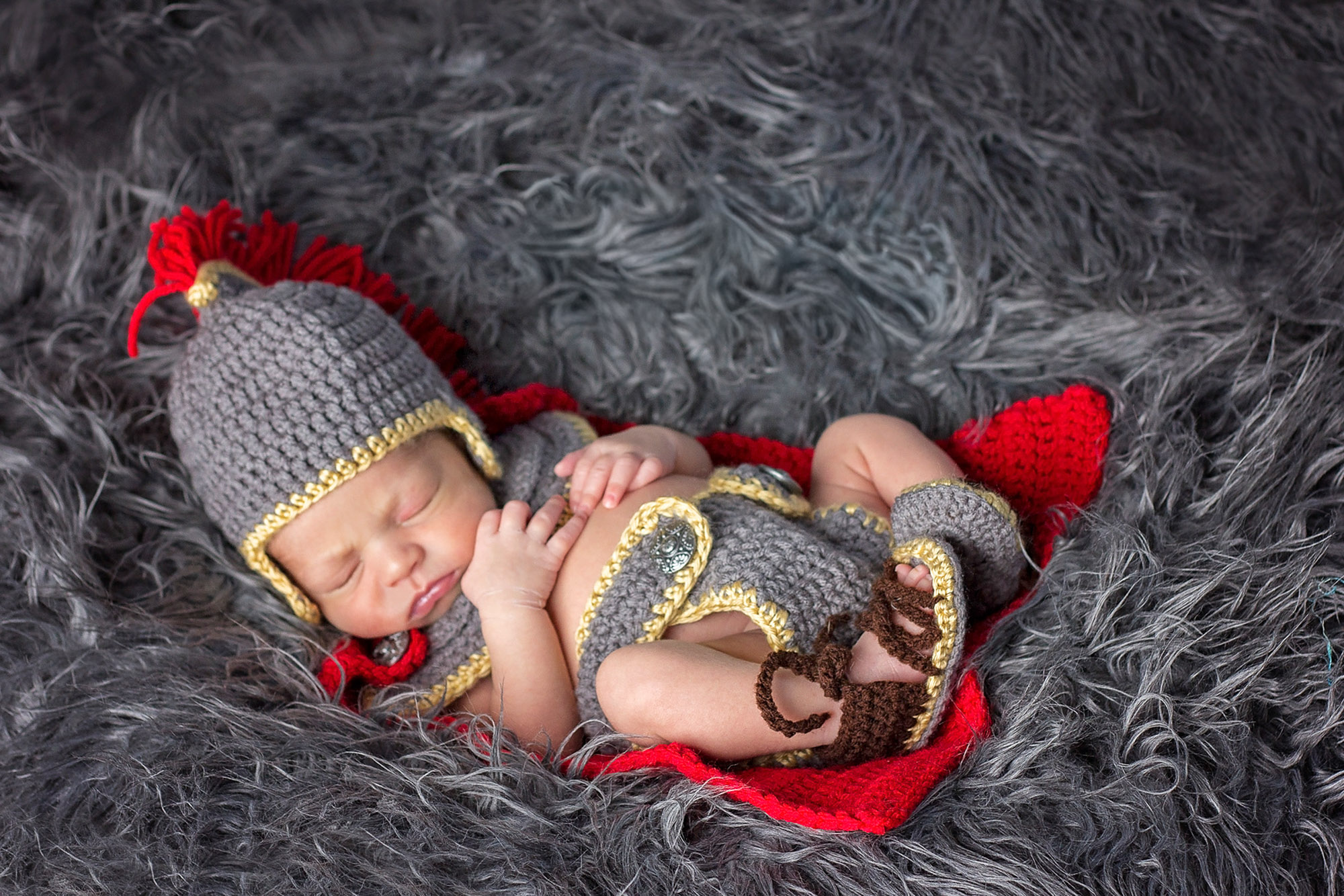 Newborn baby sleeping on gray faux fur, dressed in an adorable crocheted knight costume inspired by the chic anne hathaway crochet baby outfit, complete with a helmet, tunic, and sandals, lying on a red blanket.