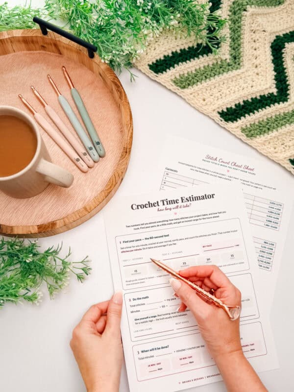 A person fills out a crochet time estimator sheet at a table with crochet hooks, a coffee cup, a tray, a green and white crochet piece, and green decorative plants.