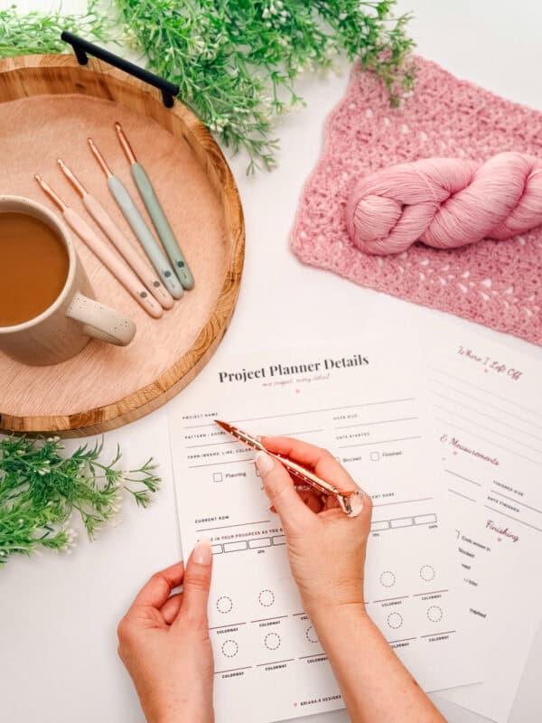 A person fills out a project planner next to a tray with coffee and crochet hooks, a pink crochet piece, yarn, and greenery on a white surface.