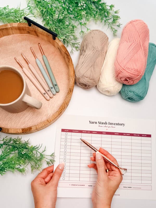 A person holds a pen and a Yarn Stash Inventory sheet next to a tray with crochet hooks, a cup of coffee, yarn skeins, and green decorative plants.