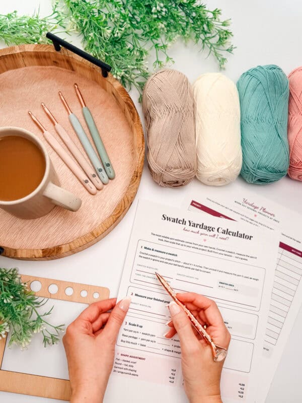 A person measures yarn with a crochet hook next to a cup of coffee, pastel yarn balls, crochet hooks, and a swatch yardage calculator worksheet on a table.