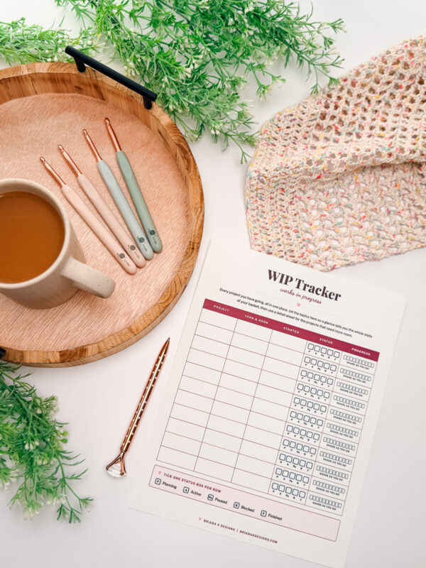 A mug, crochet hooks, a pen, a wooden tray, a crochet project, lush greenery, and a crochet WIP tracker sheet are beautifully arranged on a white surface.