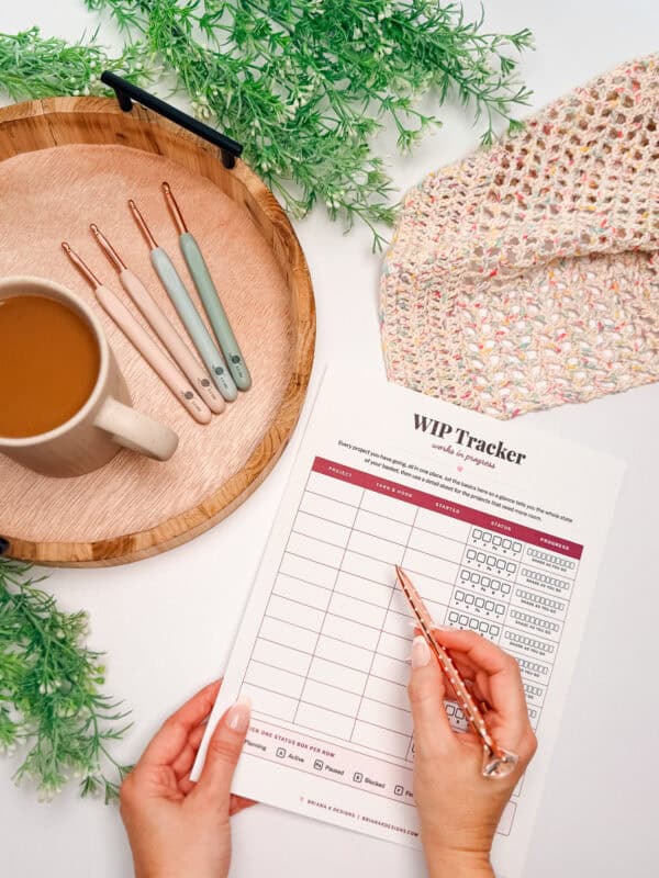 A person holds a pen above a WIP tracker sheet on a table with a mug of coffee, crochet hooks on a tray, a crochet piece, and green decorative leaves.