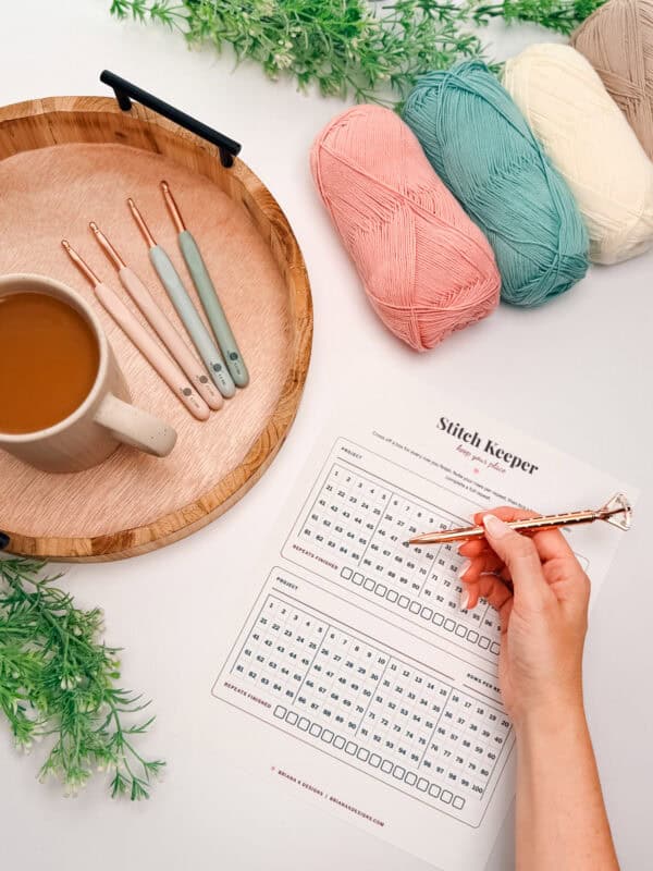 A hand holds a pen over a stitch keeper chart beside yarn balls, crochet hooks, a mug of coffee, and a wooden tray on a table with green foliage accents.