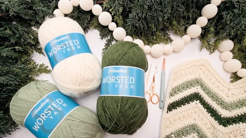 Three skeins of worsted yarn, crochet tools, a crocheted zigzag piece, wooden beads, and evergreen branches are arranged on a white surface.