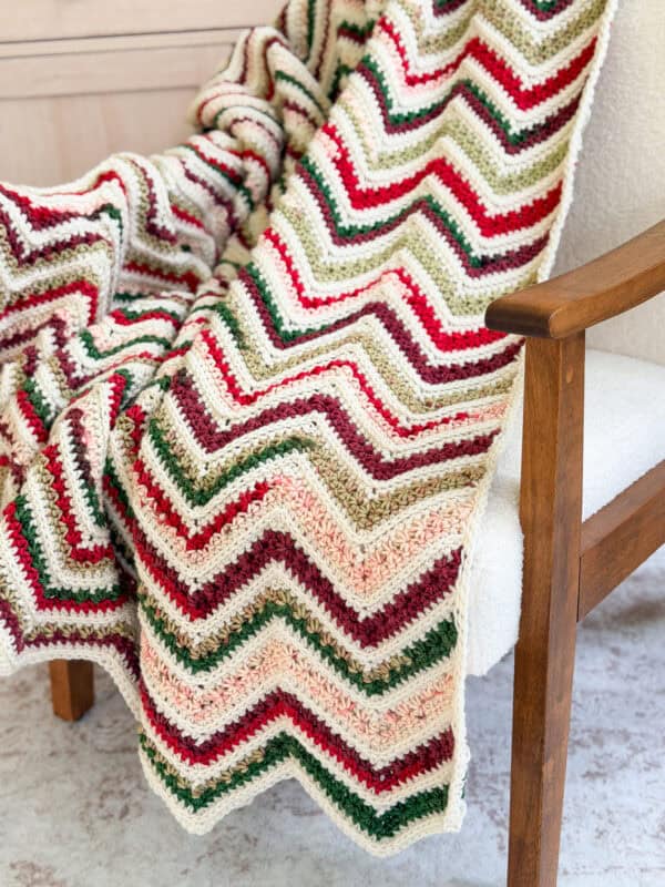 A crocheted zigzag patterned blanket in red, green, cream, and beige draped over a wooden armchair with a white cushion.