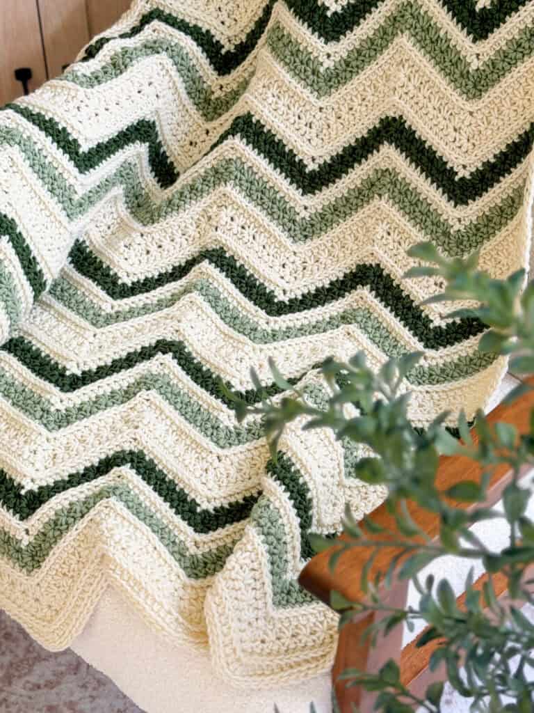 A crocheted blanket with green and cream zigzag stripes is draped over a wooden chair, with a leafy plant in the foreground.