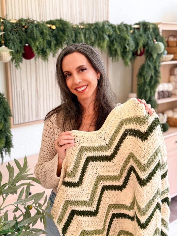 Woman smiling and holding a green and cream chevron-patterned crochet blanket; greenery and yarn shelves in the background.